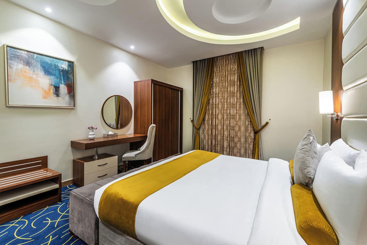 Habitación de hotel con una cama, un escritorio con silla, un armario, un espejo y un cuadro en la pared.