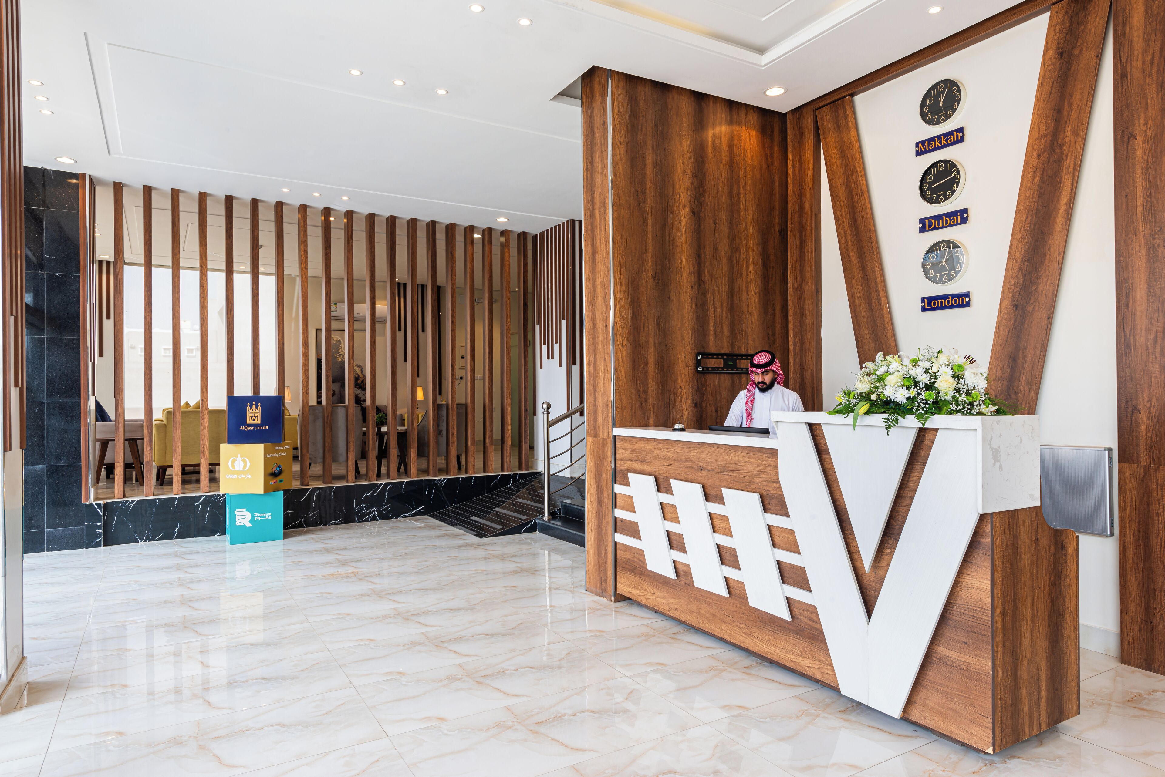 A hotel reception area with a wooden reception desk, a staircase, and a seating area with tables and chairs.