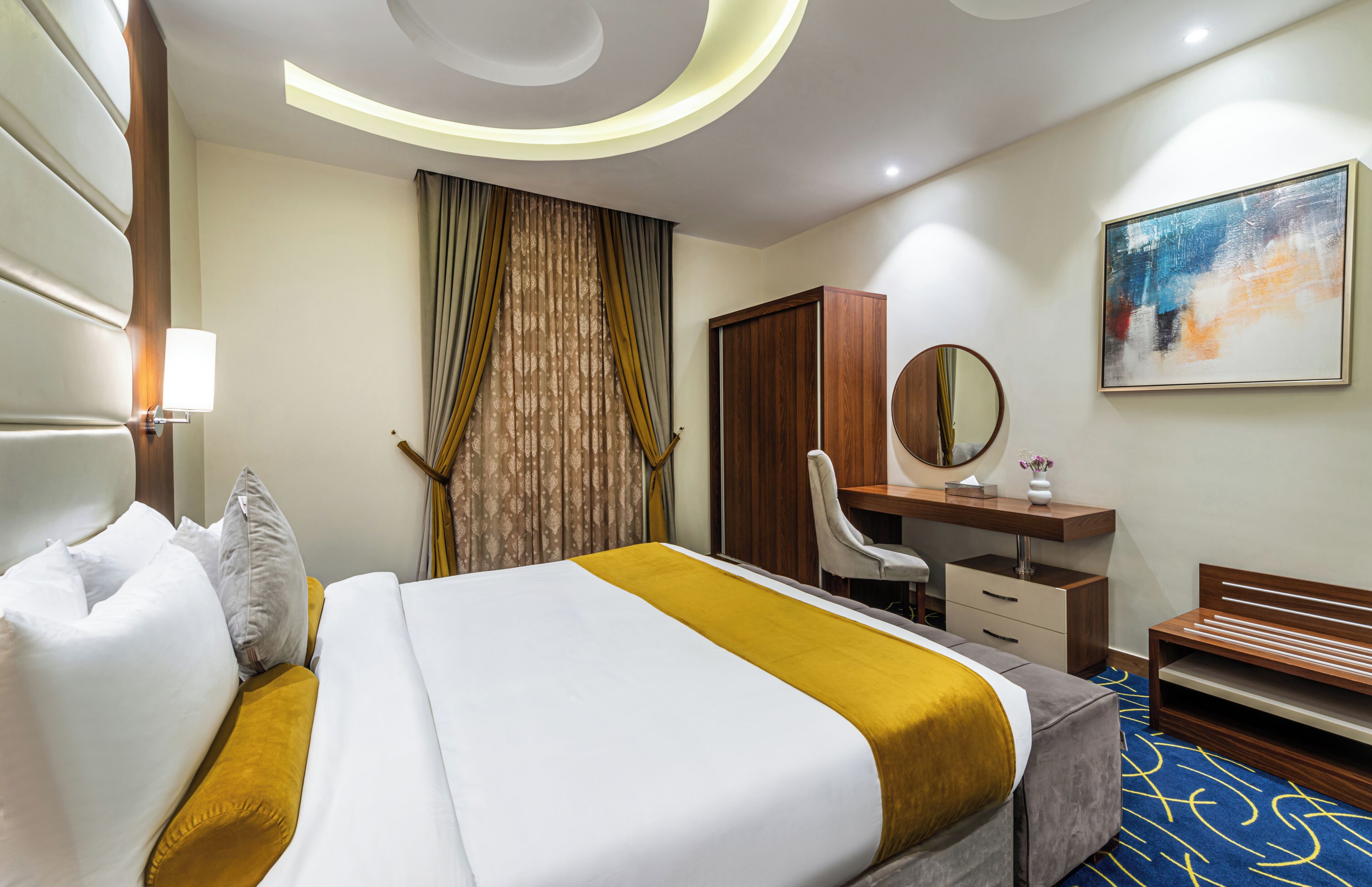 Habitación de hotel con cama, escritorio, silla, armario, espejo y un cuadro en la pared.