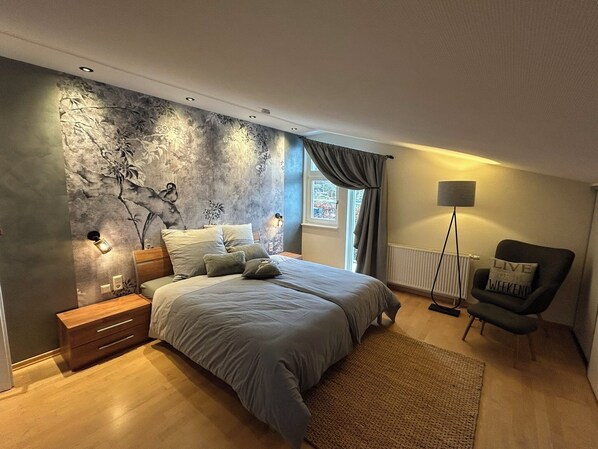 A modern bedroom with a large bed, a nightstand, a dresser, a floor lamp, and a chair. - Workshop chalet ZH10 (Olsberg)