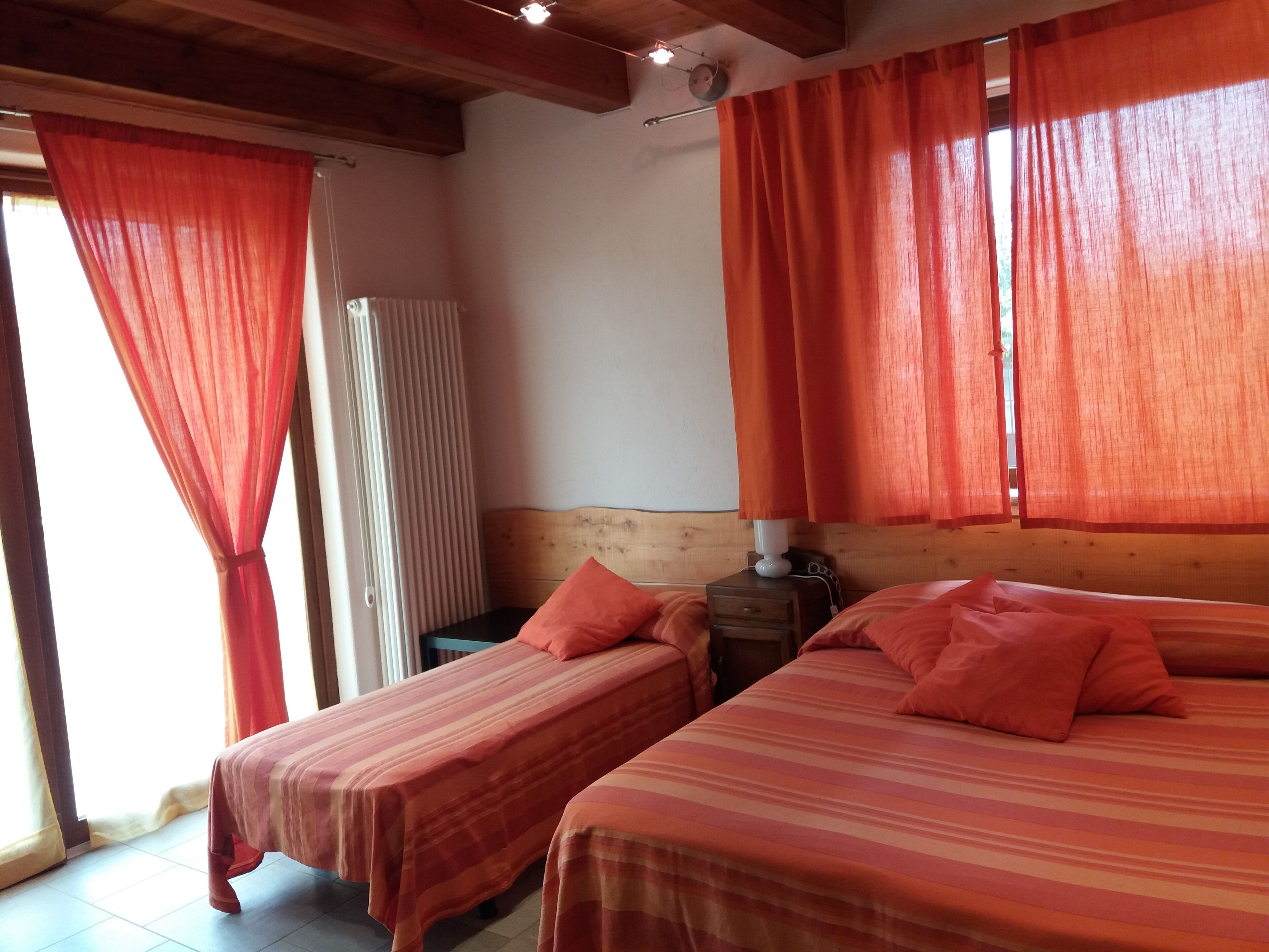 A room with two beds, each with a striped bedspread and matching pillows. There is a wooden headboard, a nightstand with a lamp, and a window with orange curtains.