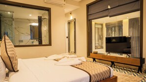 A hotel room with a large bed, a TV, a mirror, and a bathroom visible through a glass door.