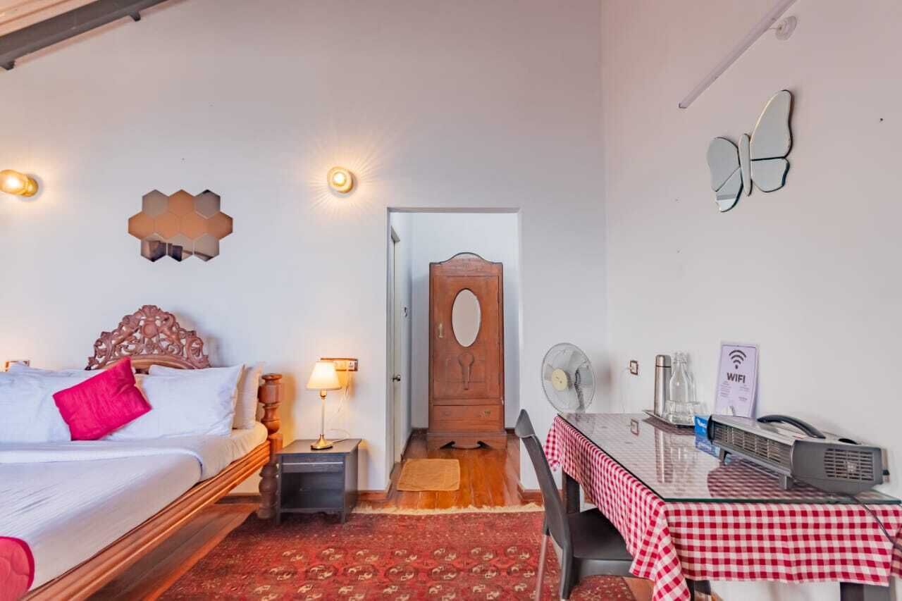A bedroom with a bed, bedside table, lamp, and a desk with a fan and a sign indicating WiFi availability.