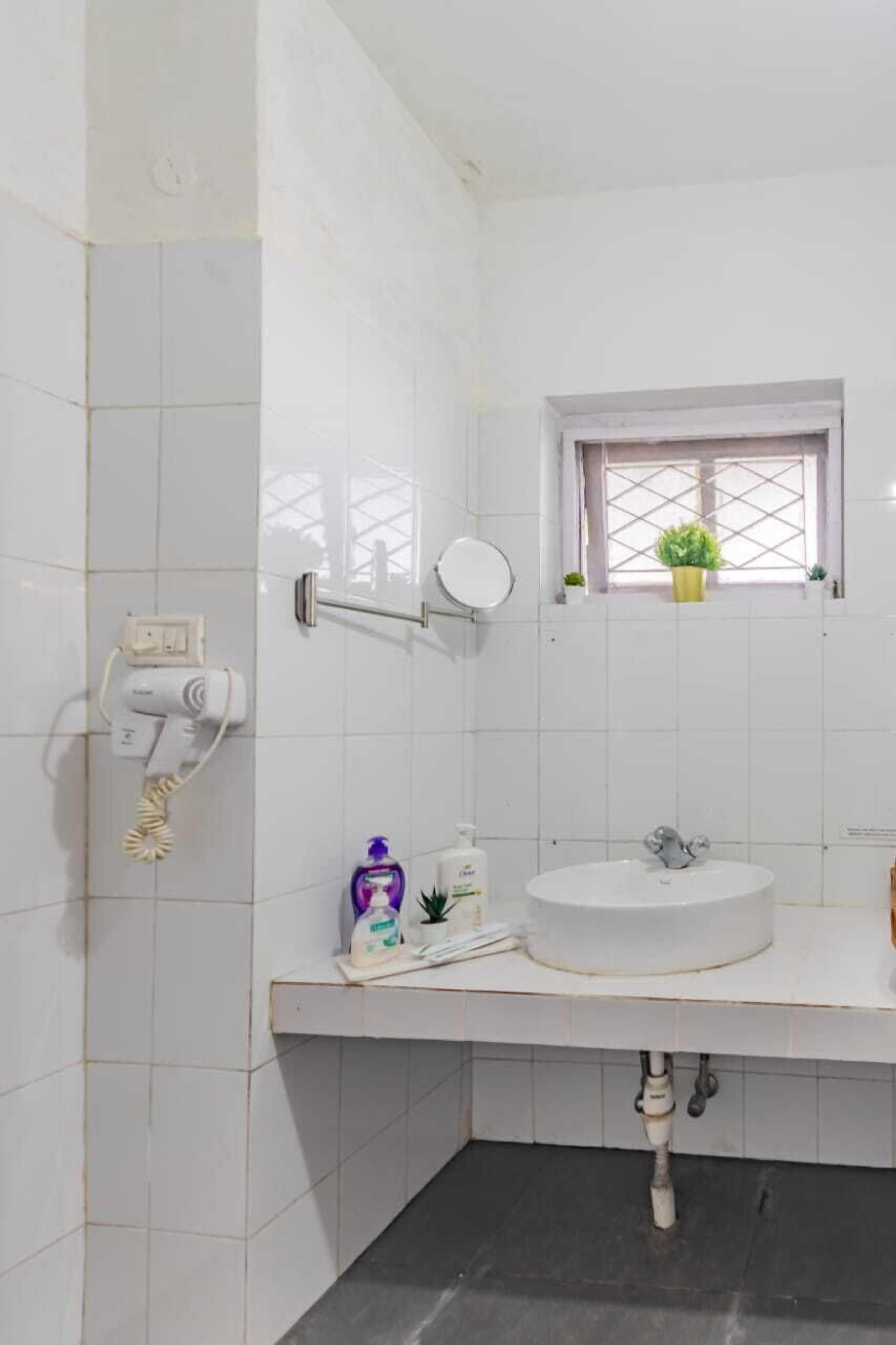 A bathroom with white tiles, a sink, a mirror, a window with plants, and a telephone.