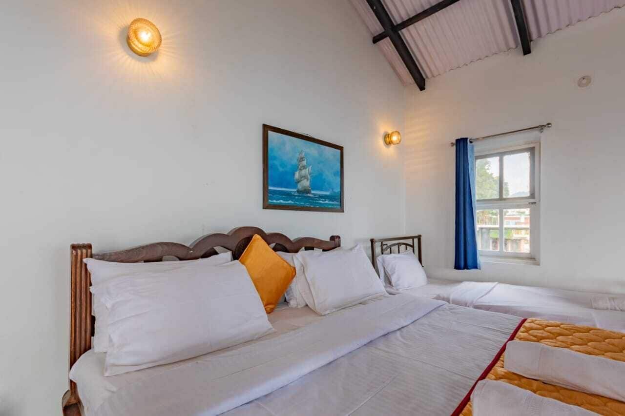 A hotel room with a bed, a window with blue curtains, a wall-mounted picture of a ship, and two wall sconces.