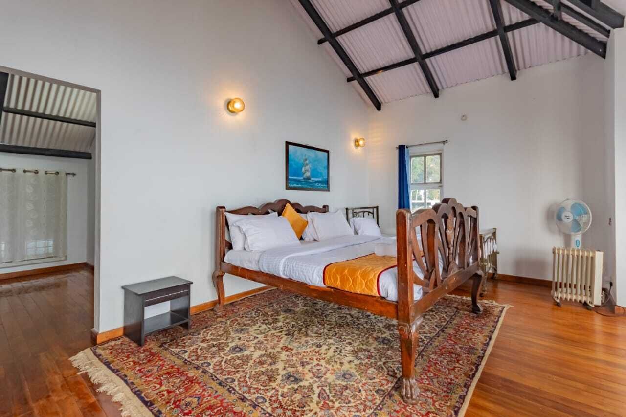 Quarto com cama de madeira, tapete estampado, mesa de cabeceira pequena, radiador e janela com cortinas.