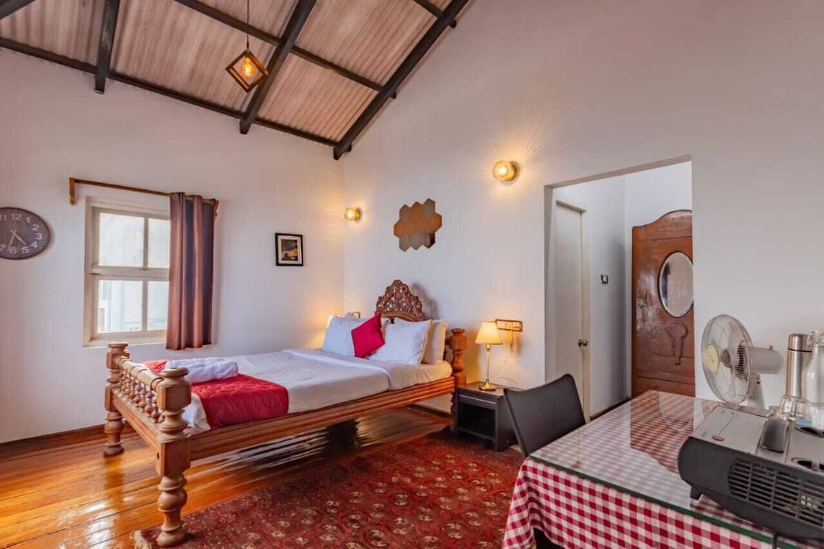 A bedroom with a wooden bed, a red and white checkered table, a clock, and a fan.