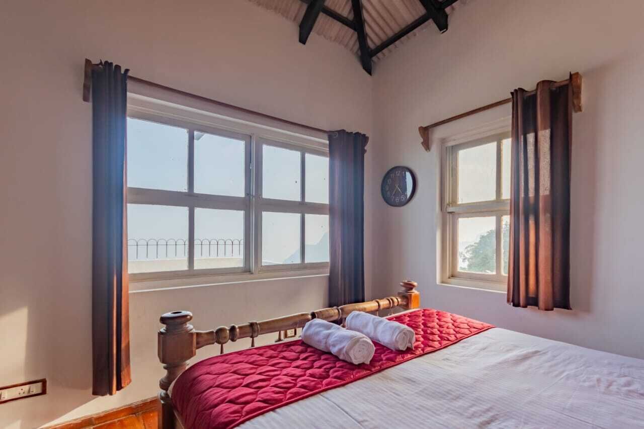 A bedroom with a bed, two windows with curtains, a clock on the wall, and a view of a balcony.