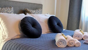 Una cama bien hecha con una colcha azul acolchada, dos almohadas decorativas negras y varios toallas enrolladas con detalles dorados.