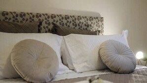 Una cama bien hecha con almohadas blancas y color beige, un cabecero estampado y una lámpara de noche.