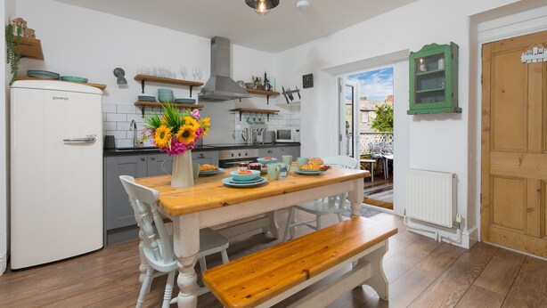 A kitchen with a wooden table, white chairs, and a bench. There is a refrigerator, a stove, and a window with a view of the outside. - Benjamin’s Bolthole, St Ives- Sleeps 4 - Outside Space – Dog-Friendly (St Ives)