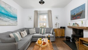 A modern living room with a grey sectional sofa, a wooden coffee table with a bottle and snacks, a fireplace, and a large painting of a boat on the wall. - Benjamin’s Bolthole, St Ives- Sleeps 4 - Outside Space – Dog-Friendly (St Ives)