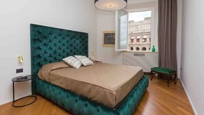 A bedroom with a teal upholstered headboard, a wooden floor, a bed with a beige mattress, a bedside table with a lamp, and a window with a view of a historical building.
