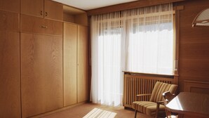 Quarto com guarda-roupa de madeira, uma cadeira, uma mesa e janela com cortinas leves.