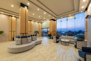 A modern hotel lobby with a curved sofa, a coffee table, and a view of the cityscape through large windows. - Al Qasr Hotel Suites - Al Iskan District (Khamis Mushait)