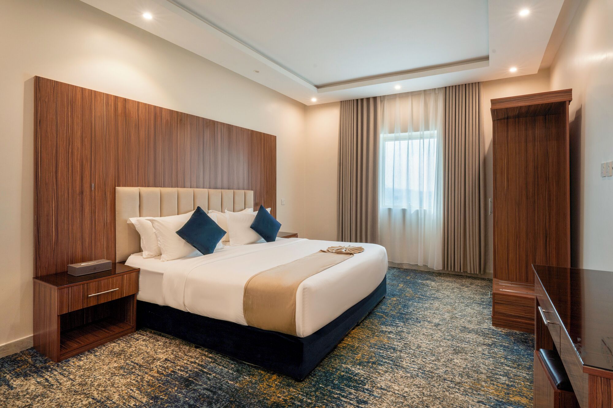 A hotel room with a large bed, wooden headboard, a bedside table, a flat-screen TV, and a carpeted floor.