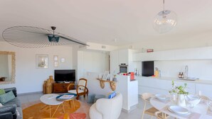 A modern living room with a sofa, coffee table, and a TV. There is a dining area with a table set for two. The kitchen is visible with white cabinets and a countertop. - NICE 130m2 Terrace with PANORAMIC VIEW, Swimming Pool, Free Parking, A/C, WIFI (Nice)