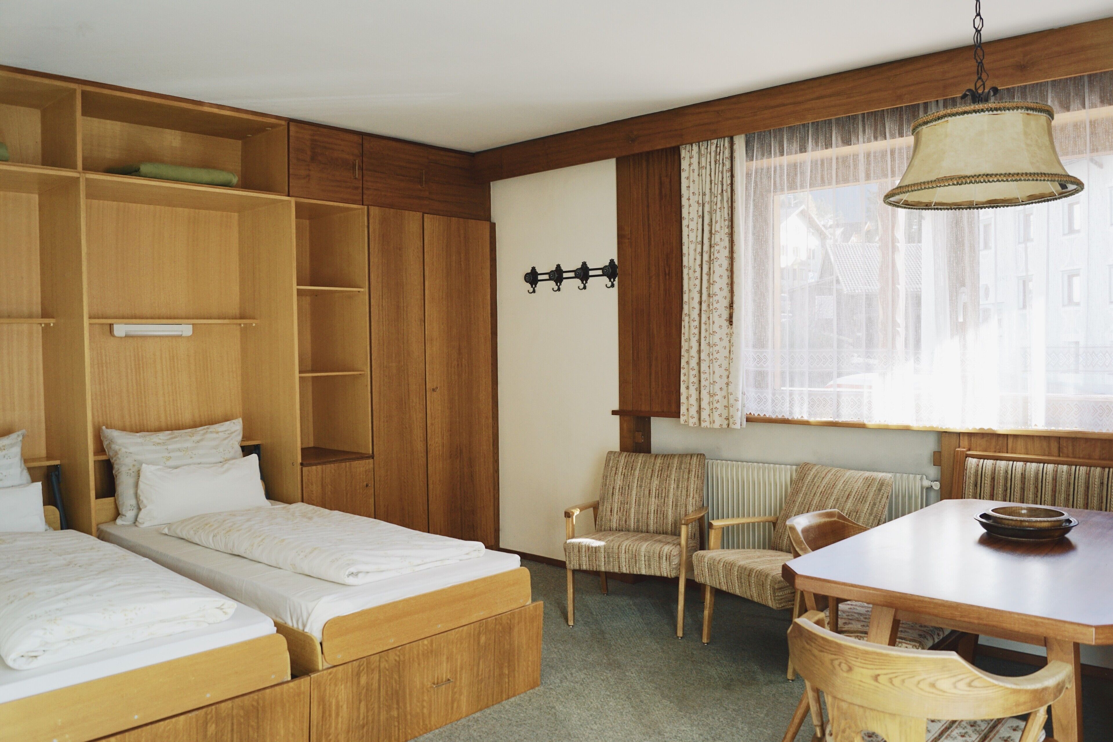 Une chambre avec deux lits, une armoire en bois, une table à manger avec des chaises et une fenêtre avec des rideaux.