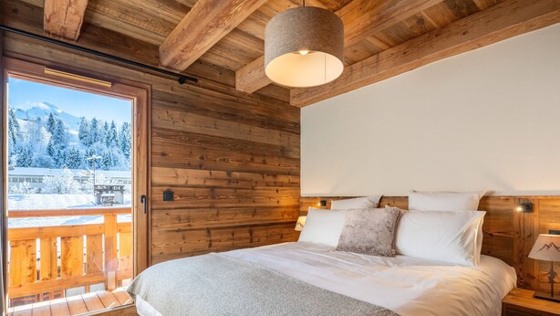 A wooden cabin bedroom with a large bed, a hanging light fixture, and a view of snow-covered mountains through a window.