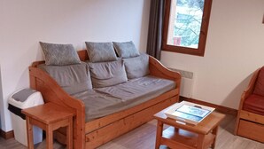 Un canapé en bois avec des coussins gris, une petite table en bois et une chaise, dans une pièce dont le sol est recouvert de planches de bois et qui possède une fenêtre.
