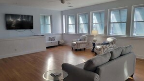 Een ruime woonkamer met houten vloeren, een grijze bank, gestreepte fauteuils, een glazen salontafel en een plafondventilator.