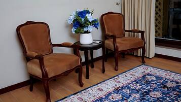 To træstole med polstrede sæder, et rundt bord med en hvid vase med blå blomster, og en tæppe med et mønster på et flisegulv.