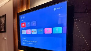 Ein wandmontierter Flachbildfernseher mit Google TV-Oberfläche, eine moderne Konsole mit Dekorationselementen und eine Wand mit Marmoroptik.