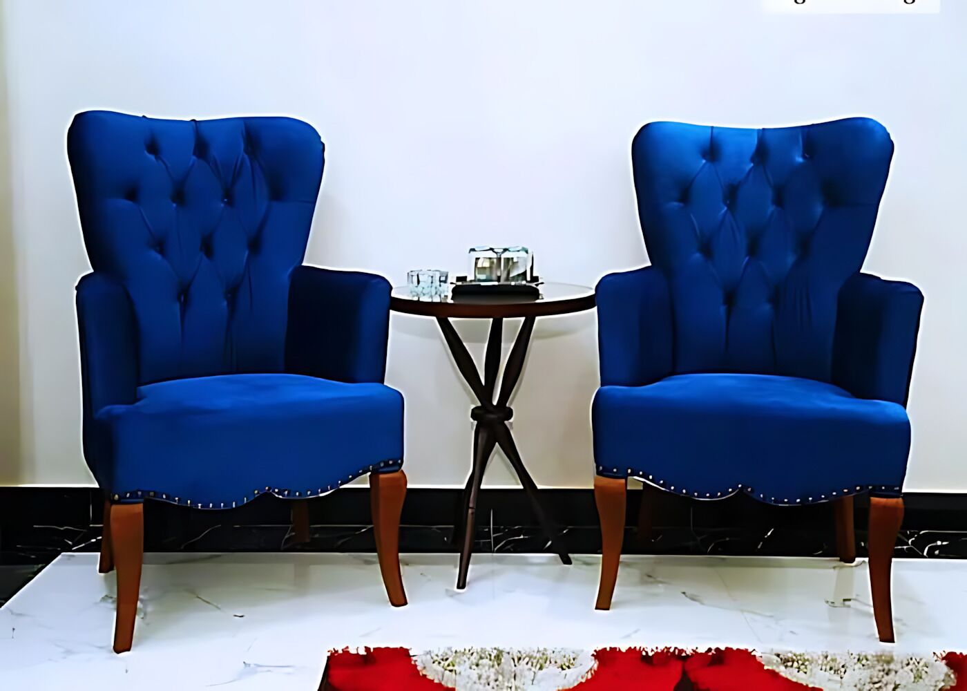 Twee blauwe fauteuils met houten poten, een kleine ronde tafel met een glazen blad, en een witte muur op de achtergrond.