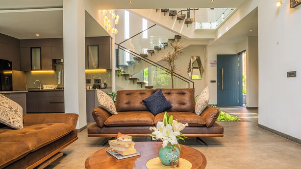 Ein modernes Wohnzimmer mit einem braunen Ledersofa, einem Couchtisch mit Büchern und einer Blumenvase, und einer Treppe, die zu einer oberen Ebene führt.