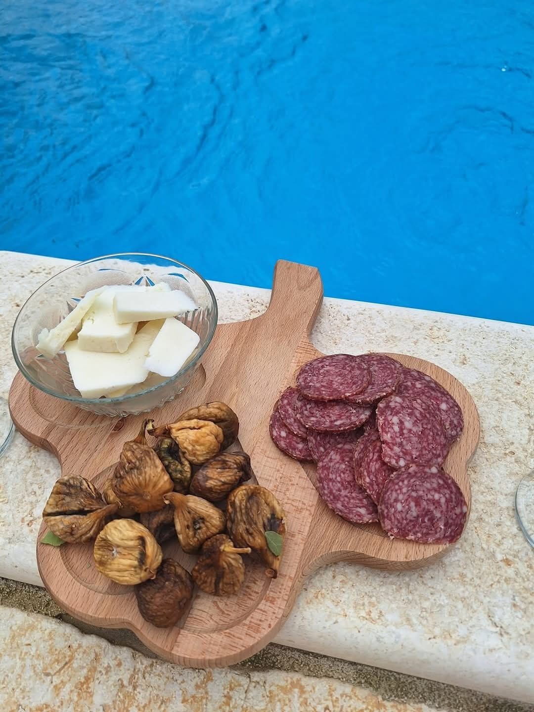 A wooden board with sliced salami, walnuts, and cheese by a poolside.