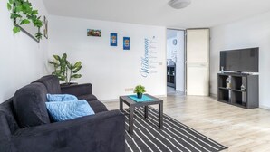 Ett modernt vardagsrum med en svart soffa, ett litet bord med en växt, en tv på ett tv-bord och en vägg med ett välkomstmeddelande.
