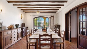 A dining area with a long table, wooden chairs, a wooden cabinet, and a view through an arched window.