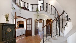 A grand staircase with a curved design, leading to a second floor. There is a bed with a white bedspread and a wooden headboard, a dark wooden cabinet, and a potted plant.
