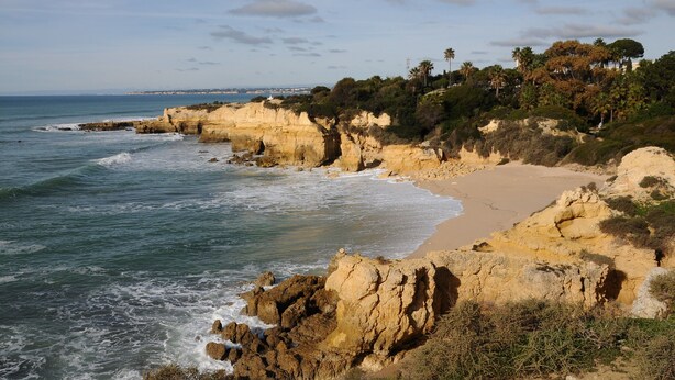 A coastal landscape with rocky cliffs, a sandy beach, and a view of the sea.