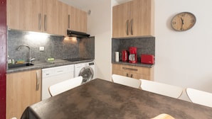 A compact kitchen with wooden cabinets, a dishwasher, a washing machine, a table with chairs, and a clock on the wall.