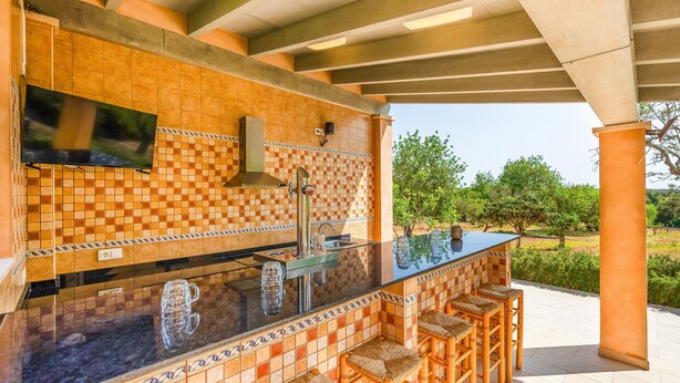 An outdoor kitchen with a tiled backsplash, a TV mounted above the counter, and a bar area with stools.