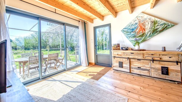 A spacious room with large sliding glass doors leading to an outdoor area, wooden flooring, a ceiling with exposed beams, and a rustic wooden cabinet with a painting.