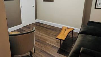 Una sala de estar con un sofá de cuero marrón oscuro, una mesa de centro de madera y una silla. Hay una puerta blanca y un cuadro en la pared.