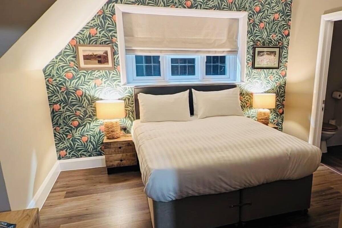 A bedroom with a bed, two bedside tables, a window with blinds, and a wall with a leafy pattern.