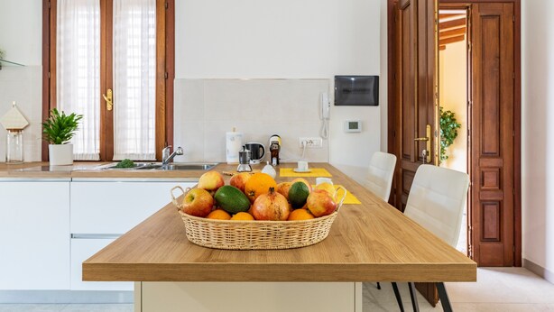 A kitchen with a wooden table, a fruit basket, a coffee machine, and a TV on the wall. - Holiday Home 'Laura' with Private Terrace, Wi-Fi and Air Conditioning (Bari Sardo)