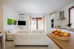 A modern living room with a sofa, a TV, a window with curtains, and a kitchen area with a fruit basket. - Holiday Home 'Laura' with Private Terrace, Wi-Fi and Air Conditioning (Bari Sardo)