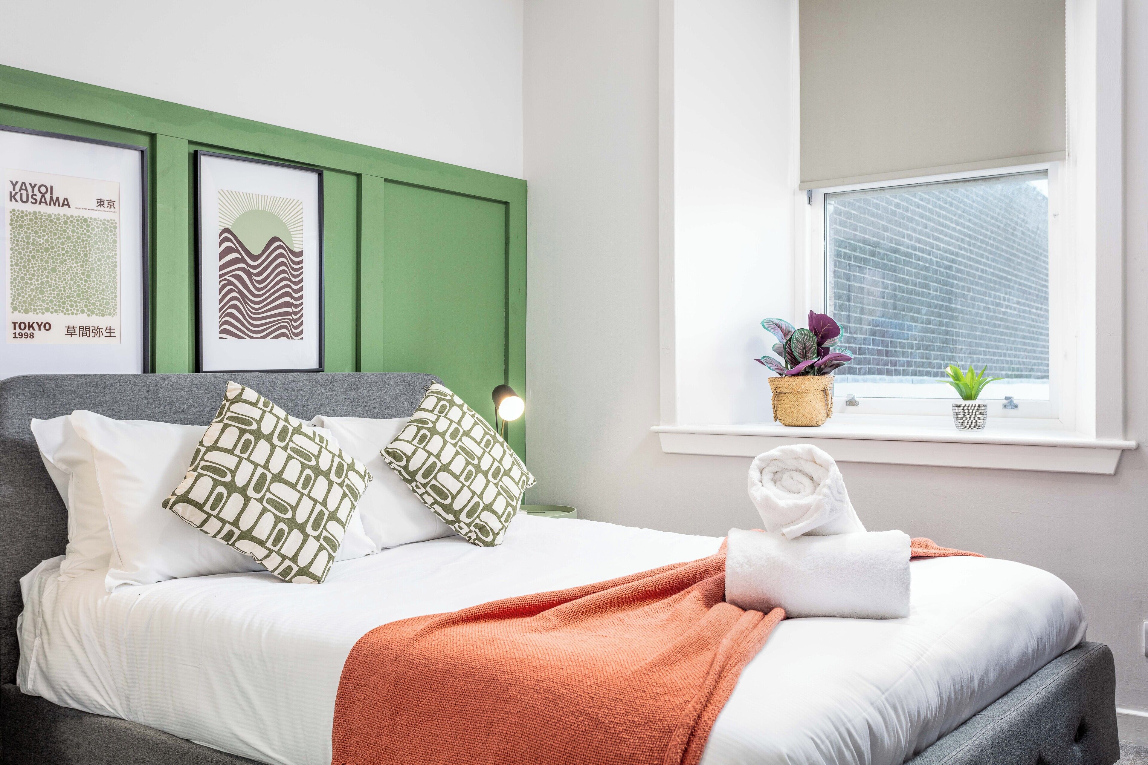 Een keurig opgemaakt bed met witte lakens en groene kussens, een groen hoofdbord en een raam met planten.