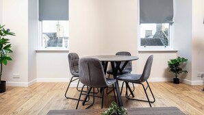 A dining area with a round table and four chairs, two windows with blinds, and potted plants. - The Annick Suite No.2 (Irvine)