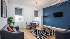 A modern living room with a blue accent wall, a round dining table, grey sofas, and a mounted TV. - The Annick Suite No.2 (Irvine)