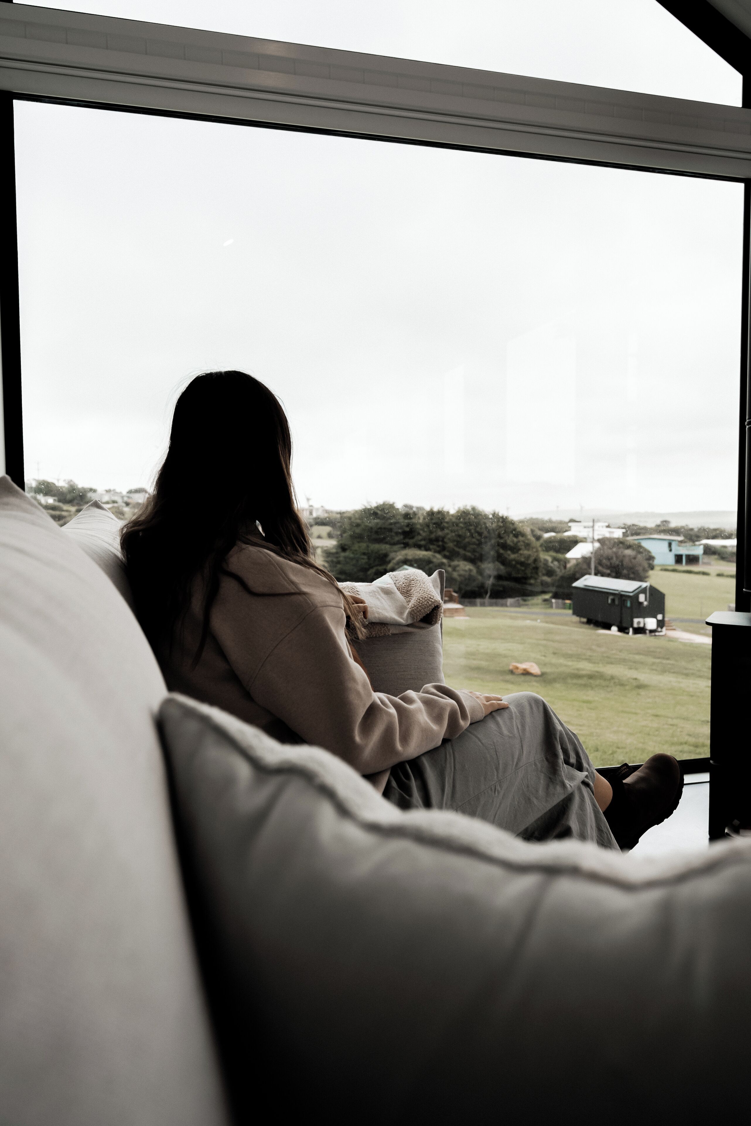 Una persona sentada en un sofá junto a un ventanal grande con vistas a un área verde y un edificio a lo lejos.