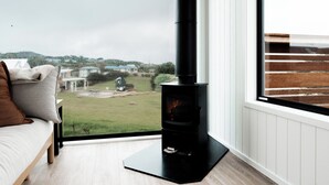 Una habitación con un ventanal grande, un horno de leña de madera negra, suelo de madera y una cama con almohadas.