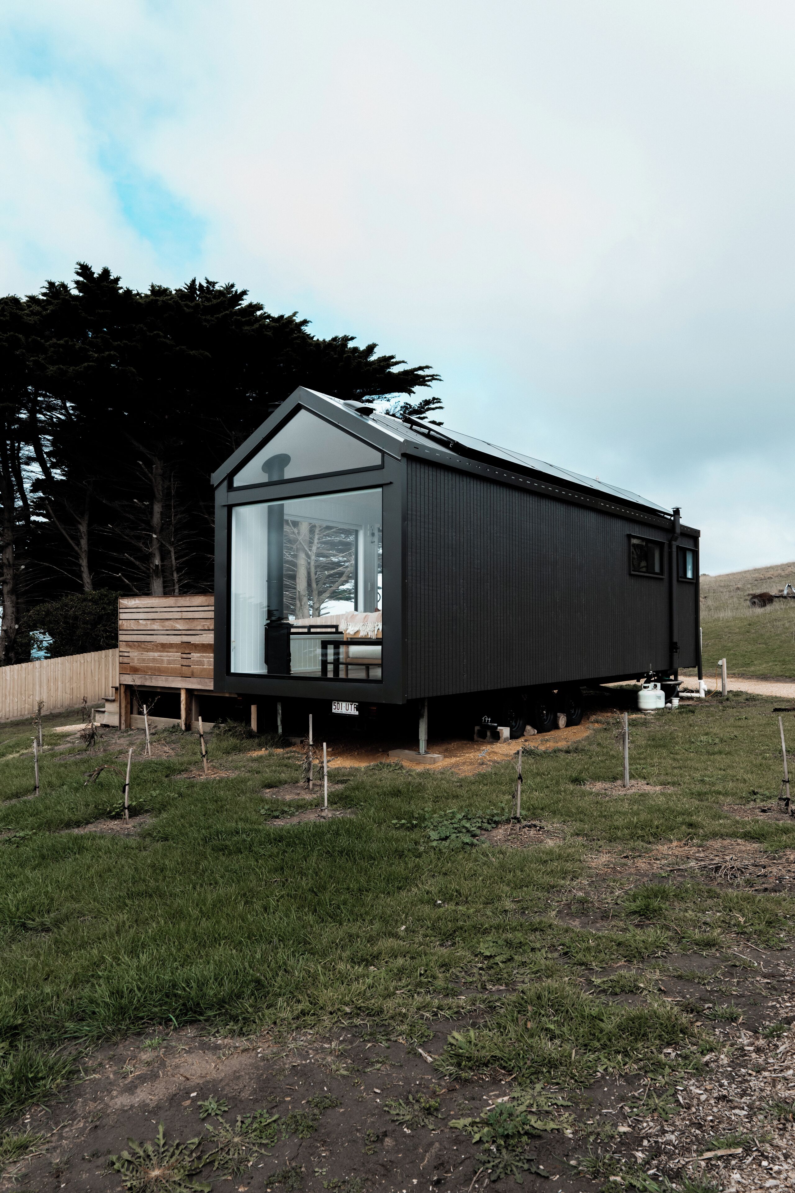 Una cabaña moderna de color oscuro con un amplio ventanal, una terraza de madera y un paisaje con césped al fondo.