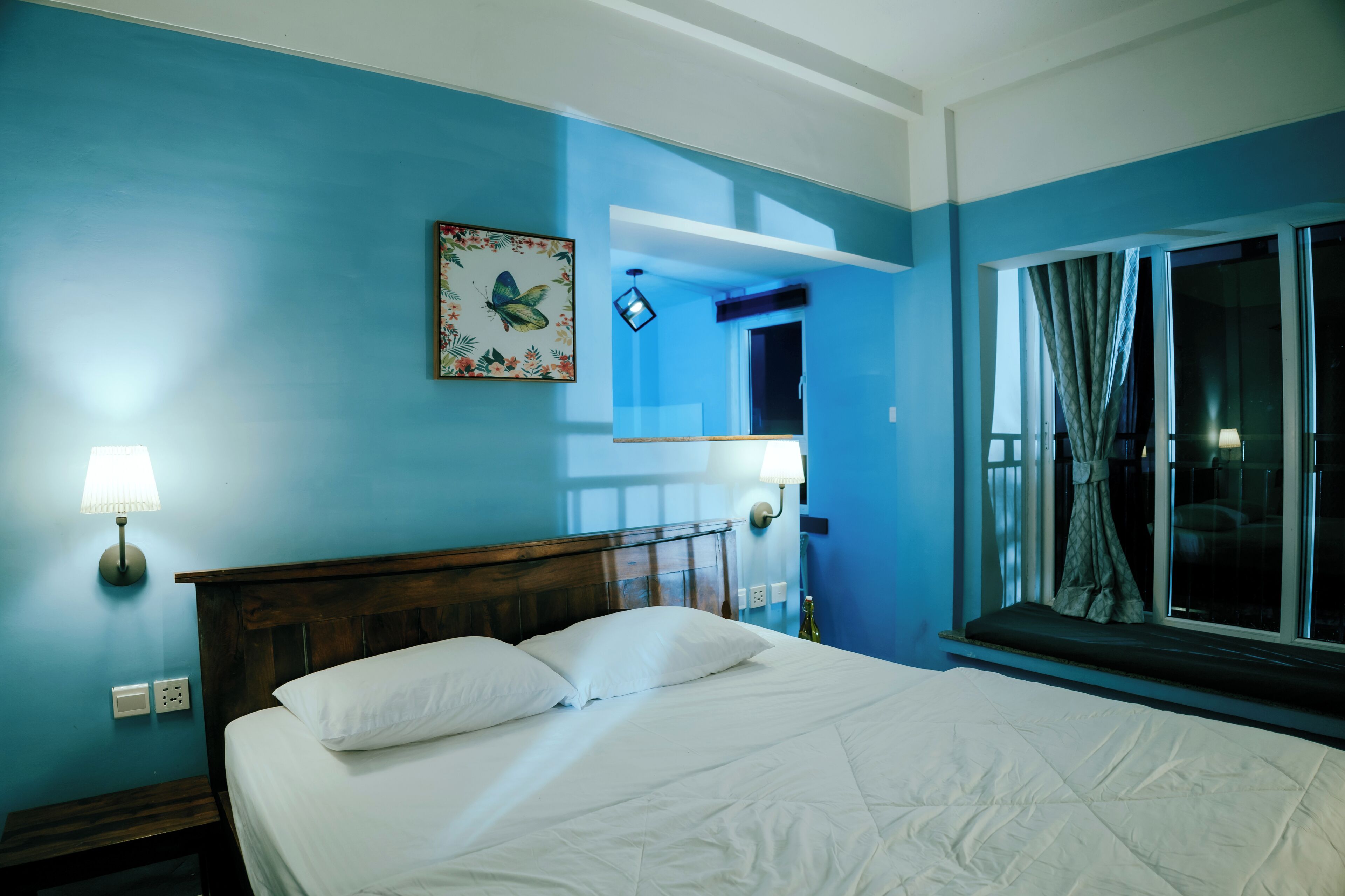 Un dormitorio con cabecera de madera, cama con ropa blanca, mesita de noche, lámpara de pared, ventana con cortinas y un espejo.