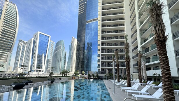 A swimming pool with lounge chairs and palm trees, surrounded by high-rise buildings. - Easygo-cozy 1bdr Zada Tower (Dubai)