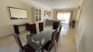 Een moderne eetruimte met een glazen tafel en stoelen, een bank op de achtergrond, en een aan de muur bevestigde televisie.